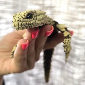 Könnte beinhalten: Eine 3D-gedruckte, bewegliche Echsenfigur mit einer texturierten, beigefarbenen, schuppigen Oberfläche und einer dunkelbraunen Unterseite. Die Echse hat einen detaillierten Kopf mit sichtbaren Augenmerkmalen und gegliederten Gliedmaßen.