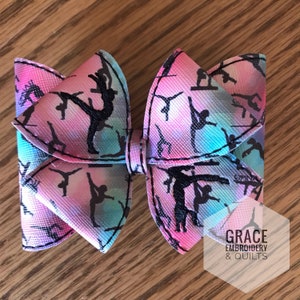 Könnte beinhalten: Eine rosa und blaue Schleife mit einem schwarz-weißen Muster von Turnerinnen. Die Schleife ist aus Stoff und hat eine Satin-Oberfläche. Der Text "GRACE EMBROIDERY & QUILTS" ist in der unteren rechten Ecke des Bildes sichtbar.