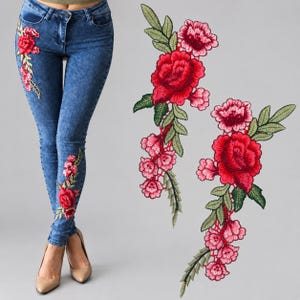 Puede incluir: Pantalones vaqueros de mezclilla azul con efecto lavado al ácido y parches florales bordados de rosas rojas y rosas con hojas verdes. El bordado se sitúa en la cadera derecha y en la parte inferior de la pierna izquierda, junto a dos parches.