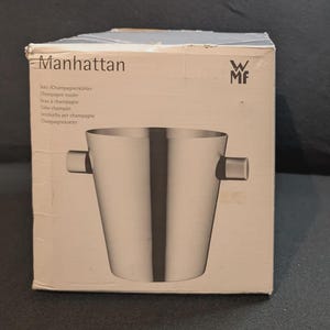 May include: A WMF Manhattan champagne cooler in a cardboard box. The box is white with the text Manhattan and the WMF logo printed on the front. An illustration of the stainless steel champagne cooler with two side handles is displayed.