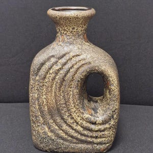 Pode incluir: Um vaso de cerâmica com um acabamento texturizado e salpicado em tons de castanho e bege. O vaso apresenta um corpo plano e retangular com cantos arredondados, um pescoço estreito e um recorte circular distinto no lado direito com relevos.