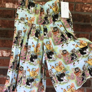 May include: Light blue culottes with a whimsical print of kittens, watering cans, flowers, and butterflies. The fabric is gathered at the waist and hangs loosely. The culottes are hanging on a clothesline with a price tag.