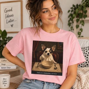 May include: A light pink t-shirt featuring a graphic print of a small dog wearing a white neckerchief, sitting at a table with a bowl, fork, and spoon. The text below the image reads, "THIS IS A PERSONAL ATTACK" in white capital letters.