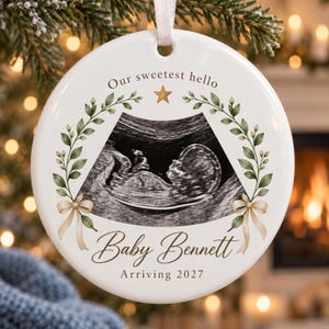 May include: A round white ceramic ornament featuring a black and white ultrasound image of a fetus. The text reads Our sweetest hello, Baby Bennett, and Arriving 2027. It is decorated with green leafy branches, a gold star, and beige ribbons.