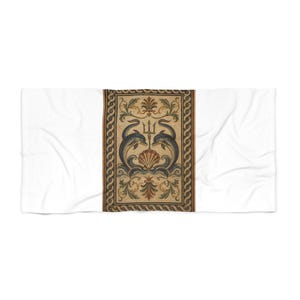 May include: A white rectangular beach towel featuring a central vertical panel with a classical mosaic-style design. The print displays two dark blue dolphins flanking a central trident and seashell, surrounded by ornate floral scrolls and a rope border.