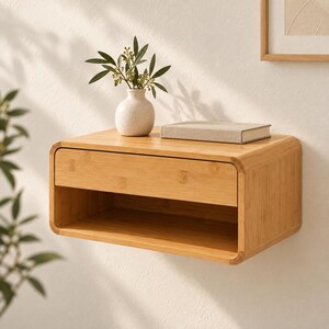 May include: A light-toned wooden floating nightstand with rounded corners, featuring a single drawer and an open storage shelf underneath. A small white ceramic vase with green foliage and a closed book are placed on the top surface.
