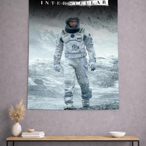 Puede incluir: Un tapiz de pared rectangular que presenta el póster de la película Interstellar. La imagen muestra a un astronauta con un traje espacial blanco de pie sobre un paisaje nevado y helado. La palabra INTERSTELLAR está impresa en blanco arriba.