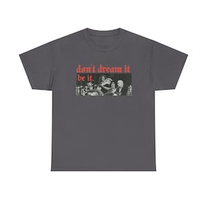May include: A charcoal grey short-sleeve t-shirt featuring a black and white photographic print of a group of people. Above the image, the text "don't dream it" is printed in red, and below the image, the text "be it." is printed in red.
