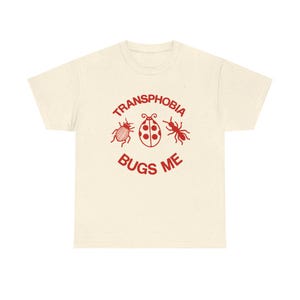Può includere: Una t-shirt a maniche corte color crema con una stampa grafica rossa. Il design include tre illustrazioni di insetti, uno scarabeo, una coccinella e una formica, tra il testo rosso "TRANSPHOBIA BUGS ME" disposto in modo circolare.
