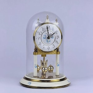 Puede incluir: Un reloj de cuarzo tipo aniversario con esfera blanca, números arábigos negros y detalles florales azules, bajo una cúpula de cristal. El reloj presenta un marco de metal dorado, pilares blancos con motivos florales y un péndulo giratorio.