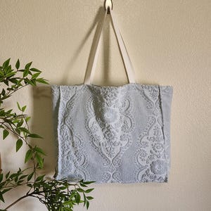 May include: A light blue tote bag made from textured terry cloth fabric featuring a raised damask floral pattern. The bag has two long, flat, off-white fabric straps attached to the top edge, allowing it to hang vertically from a wall hook.