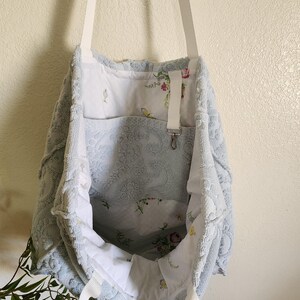 May include: A light blue textured tote bag with white fabric straps. The exterior features a raised floral pattern. The interior is lined with white fabric decorated with a colorful floral print and includes a white strap with a metal swivel clasp.
