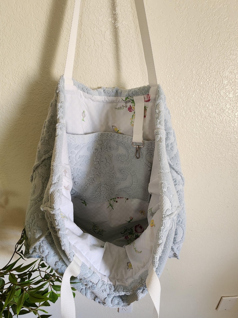 May include: A light blue textured tote bag with white fabric straps. The exterior features a raised floral pattern. The interior is lined with white fabric decorated with a colorful floral print and includes a white strap with a metal swivel clasp.