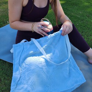 May include: A light blue tote bag made of textured terry cloth fabric featuring a raised paisley pattern and two white fabric shoulder straps. The bag has a rectangular shape and is constructed from a soft, absorbent material with a visible weave.