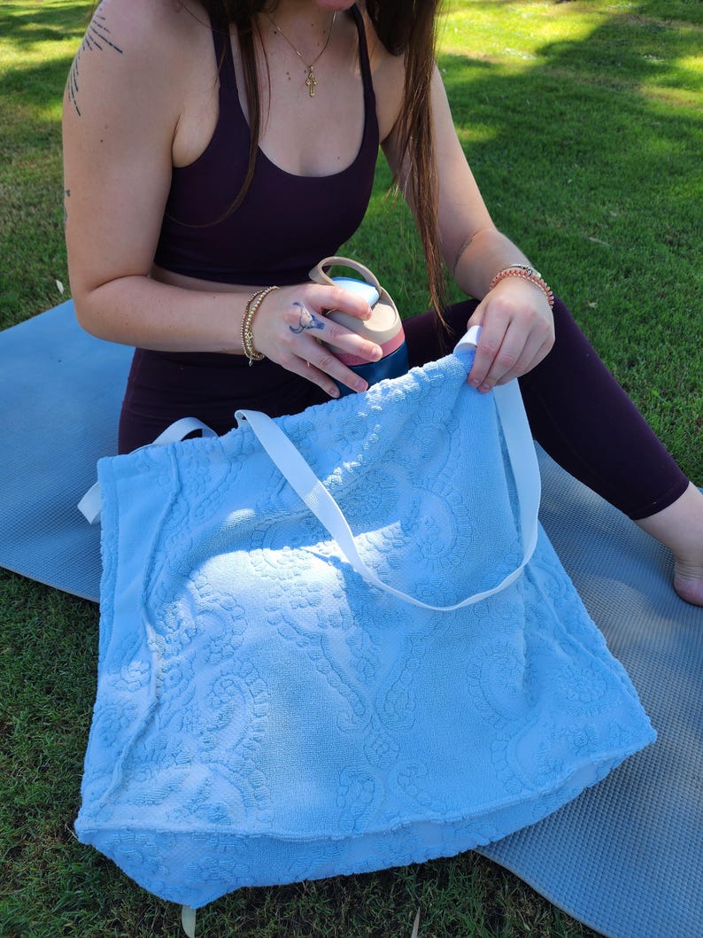 May include: A light blue tote bag made of textured terry cloth fabric featuring a raised paisley pattern and two white fabric shoulder straps. The bag has a rectangular shape and is constructed from a soft, absorbent material with a visible weave.