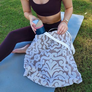 May include: A large tote bag featuring a beige and white damask pattern with white fabric straps. The bag is made of a textured material and contains a dark blue water bottle with a light blue and pink lid, which is being held by the top.
