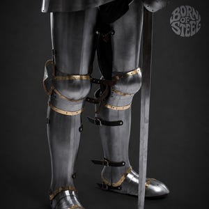 May include: A set of polished steel leg armor featuring articulated knee plates and greaves with brass trim accents. The armor includes black leather straps and buckles, and a long steel sword stands vertically beside the leg pieces. The text BORN OF STEEL is visible.