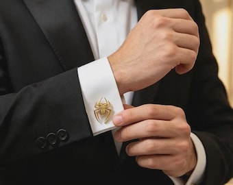 Op de afbeelding: Een goudkleurige manchetknop in de vorm van een spin, bevestigd aan de witte manchet van een overhemd. Het overhemd wordt gedragen onder een zwart colbert, waarbij een zwarte vlinderdas bij de kraag zichtbaar is. Het ontwerp toont een spinnenfiguur.