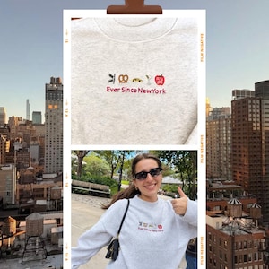 May include: A light gray crewneck sweatshirt featuring embroidered icons of a street sign, pretzel, taxi, martini glass, and a red apple, with the red text "Ever Since New York" underneath. The design is centered on the chest of the garment.