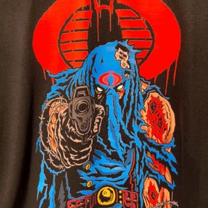 May include: A black t-shirt featuring a graphic print of a hooded figure in a blue tattered cloak, holding a handgun. The figure has a red Cobra logo on the forehead and is surrounded by barbed wire. A large red circular Cobra emblem is in the background.