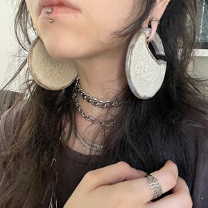 May include: Large, circular, ceramic hoop earrings with a textured, off-white, crackled glaze finish and a smooth, grey-toned outer rim. The earrings feature a distinct gap at the top where they connect to the earlobe through a gauge piercing.