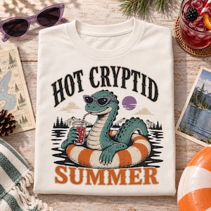 May include: A cream-colored t-shirt featuring a graphic of a green creature wearing sunglasses, floating in an orange and white striped inner tube on water, holding a red drink with a straw, with the text HOT CRYPTID SUMMER printed in black and orange.