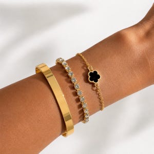 Puede incluir: Un conjunto de tres pulseras doradas. Una es un brazalete rígido de metal liso. La segunda es una cadena flexible con piedras transparentes de corte redondo. La tercera es una cadena fina con un pequeño dije negro en forma de trébol.