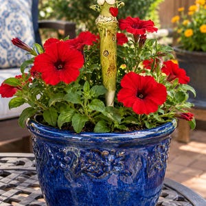 May include: A deep blue ceramic planter with an embossed floral pattern contains vibrant red petunia flowers and a whimsical light green ceramic garden stake featuring a small red heart and a cartoonish face with wide eyes.