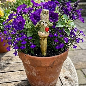 May include: A terracotta flower pot filled with vibrant purple petunias and lobelia flowers. Centered in the soil is a light green ceramic garden stake shaped like a textured tree trunk with two wide eyes and a small, bright red heart accent.