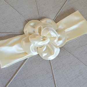May include: Ivory satin headband with a large, layered flower accent. The flower is made of the same satin material as the headband. The headband is wide and the flower is centered. The background is a light gray fabric.