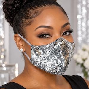May include: A face mask covered in small, reflective silver sequins. The mask features a contoured shape that covers the nose and mouth, secured by thin, light gray elastic ear loops with adjustable sliders on each side for a custom fit.