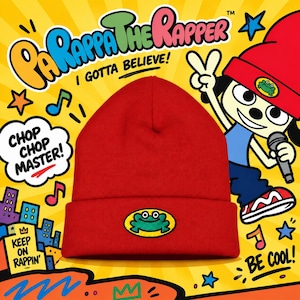 May include: A bright red beanie featuring a yellow oval patch with a green frog illustration on the front. The background displays the PaRappa the Rapper logo, the text I GOTTA BELIEVE, CHOP CHOP MASTER, KEEP ON RAPPIN, and BE COOL with cartoon graphics.