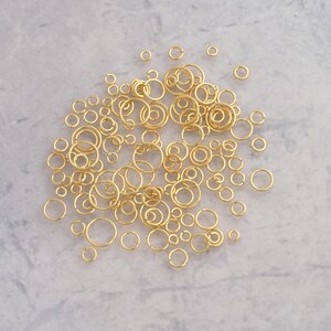 May include: A collection of numerous gold-colored metal jump rings in various sizes. Each ring features a small opening, allowing them to be used as connectors for jewelry making. The rings are scattered in a loose pile, displaying different diameters.