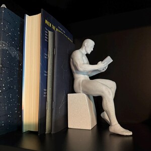 May include: A light grey, speckled, matte-finish figurine of a seated person reading a small book. The figure is positioned in a profile view, resting on a rectangular block, and is placed next to several upright books on a dark shelf.