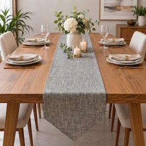May include: A long, rectangular gray table runner with a textured, woven fabric appearance centered on a wooden dining table. The runner features a pointed end and is decorated with a white floral centerpiece, two glass candles, and place settings.
