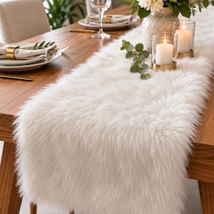 May include: A long, rectangular table runner made of white faux fur with a thick, shaggy texture. The runner is positioned lengthwise down the center of a wooden dining table, providing a soft, plush surface for decorative items and place settings.