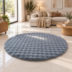 May include: A circular, dusty blue area rug featuring a textured, tufted surface with a repeating dimpled pattern. The rug has a thick, plush pile and is positioned flat on a light-colored tiled floor, showcasing a soft, uniform color throughout.