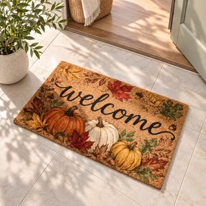 Può includere: Uno zerbino rettangolare in fibra di cocco con la scritta welcome in corsivo nero. Il design presenta tre zucche nei colori arancione, bianco e giallo, circondate da un motivo di foglie autunnali nelle tonalità del rosso, oro e verde.