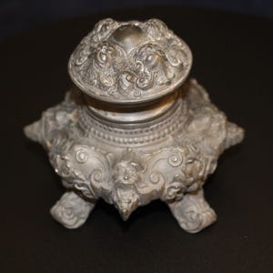 May include: A silver-toned metal inkwell featuring an ornate, embossed surface with intricate scrollwork and floral motifs. The vessel has a rounded, removable lid with matching decorative patterns and stands on four small, sculpted feet.