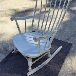 May include: A wooden rocking chair with a distressed white painted finish. The chair features a high backrest with vertical spindles, curved armrests, a smooth seat, and two curved rockers at the base. The paint shows wear along the edges.