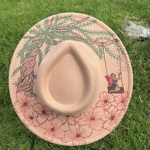 May include: A beige felt hat featuring detailed embroidery on the brim. The design includes a large green willow tree, a small figure with pink wings sitting on a swing hanging from the branches, and a cluster of pink flowers along the bottom edge.