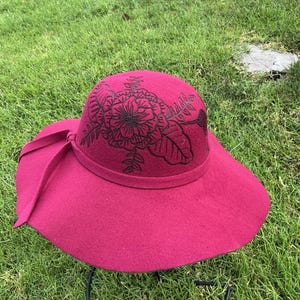 May include: A magenta wide-brimmed hat featuring black embroidered floral and leaf patterns on the crown. The hat includes a matching magenta ribbon band tied in a bow on the side, with a structured dome and a broad, slightly curved brim.