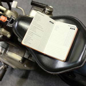 Puede incluir: Una pequeña libreta de cuero negro con una encuadernación naranja y el texto "SERVICE SCHEDULE" en la portada. La libreta está abierta en una página con una lista de verificación para el mantenimiento de la motocicleta.