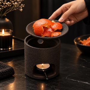 May include: A dark grey, cylindrical ceramic oil burner with a textured, stone-like finish. The burner features a circular opening at the base for a tea light candle and a removable top dish holding bright orange, fresh citrus peel pieces.