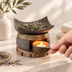 Può includere: Un bruciatore di oli essenziali in ceramica con finitura scura e testurizzata e una fascia color oro. La ciotola superiore contiene lavanda essiccata, mentre una piccola tazza abbinata con manico e lumino acceso è inserita nella base.