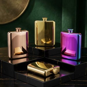 Könnte beinhalten: Vier rechteckige Metallflachmänner mit abgerundeten Ecken und Schraubverschlüssen. Ein Flachmann ist roségold, einer poliertes Gold, einer schillernd violett-blau und einer liegt horizontal in Gold. Jeder Flachmann hat eine glatte, reflektierende Oberfläche.