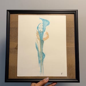 Peut inclure: Une œuvre d'art encadrée présentant un motif abstrait vertical dans des tons de bleu et d'orange clair sur fond blanc. La pièce est insérée dans un cadre noir avec un passe-partout en carton brun texturé, avec une signature dans le coin.