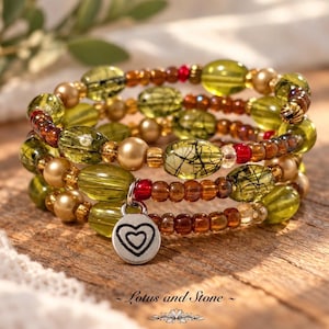 May include: A multi-strand wrap bracelet featuring olive green oval beads, metallic gold spheres, translucent brown seed beads, and small red accents. A circular silver-toned charm with an engraved heart design hangs from the bracelet.