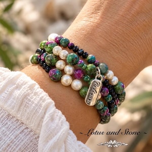 May include: A multi-strand beaded bracelet featuring a mix of round green and magenta speckled stone beads, smooth white pearls, and small black beads. A silver-toned metal charm hangs from the bracelet, engraved with the word Believe.
