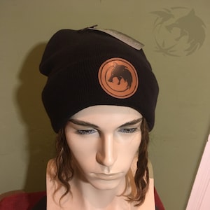 May include: Black beanie with a brown leather patch featuring a wolf design.